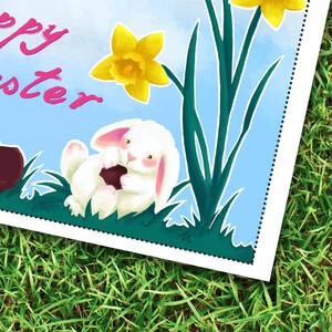 May include: A white bunny rabbit holding a chocolate egg sits in a field of green grass with yellow daffodils and the words "Happy Easter" in pink letters.