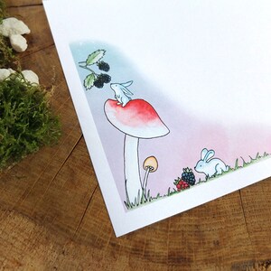 May include: A watercolor illustration of a white rabbit sitting on top of a red and white mushroom with a green stem. There are blackberries on a branch above the rabbit and another white rabbit in the grass below the mushroom.