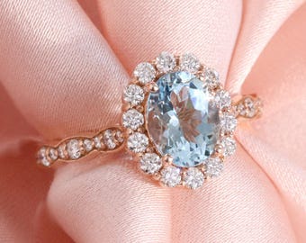May include: A rose gold ring with an oval, light blue gemstone surrounded by a halo of smaller, clear stones. The ring's band features a delicate, repeating pattern. The ring is displayed on a soft, pink fabric.