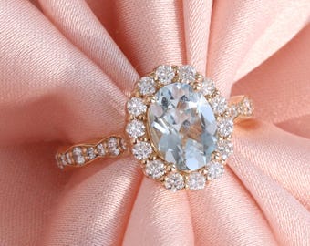 May include: A gold ring with an oval, light blue gemstone surrounded by small, clear gemstones. The ring's band features a delicate design with additional small, clear gemstones. The ring is set against a soft, pink fabric.