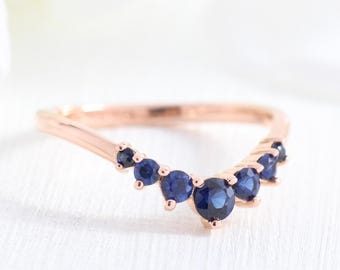 May include: Rose gold ring with a curved design. The ring features a series of round, deep blue gemstones set along the curve. The gemstones are of varying sizes, creating a delicate and elegant look. The ring is set against a white background.