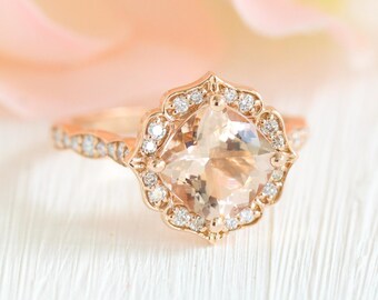 Könnte beinhalten: Ein roségoldener Verlobungsring mit einem quadratischen, pfirsichfarbenen Mittelstein. Der Ring ist von einem Kranz aus kleinen, klaren Diamanten umgeben und hat ein zartes, verziertes Band. Der Ring liegt auf einer weißen Oberfläche mit einem weichen, verschwommenen Hintergrund.