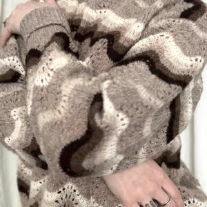May include: A cozy, long-sleeved sweater in shades of brown, beige, and white, featuring a wave-like pattern. The sweater has a high neckline and a textured knit design, perfect for cooler weather.