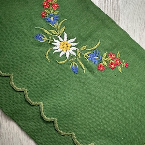Può includere: Tovaglietta da tavola in tessuto verde con un ricamo di fiori bianchi, gialli, blu e rossi. La tovaglietta ha un bordo a festoni.