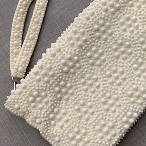 May include: A white beaded clutch bag with a matching beaded strap. The bag is made of small, round beads that are sewn together to create a textured surface.