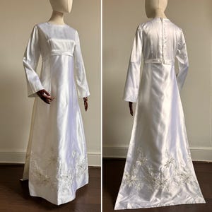 Puede incluir: Un vestido de novia de satén blanco con mangas largas y un corpiño ajustado. El vestido presenta un delicado bordado floral a lo largo del dobladillo.