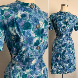 May include: A vintage dress featuring a floral pattern in shades of blue, teal, and purple. The dress has short sleeves, a high neckline, and a button-down front. The waist is cinched with a matching belt, and the skirt falls to the knee.
