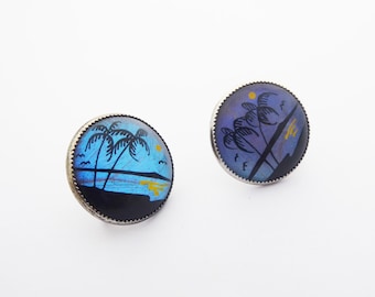 May include: A pair of round, silver earrings featuring blue and purple butterfly wings with a painted scene of palm trees and a sunset.