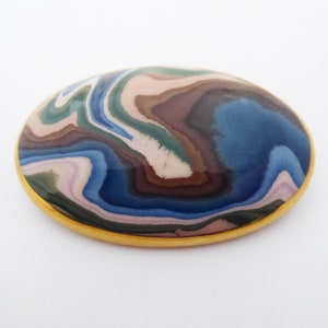 May include: A round, oval-shaped brooch with a gold rim. The brooch features a swirling pattern of blue, brown, pink, and green colours.