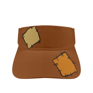 May include: A brown sun visor featuring a printed design of two square patches. One patch is light beige and the other is a darker orange, both outlined with black stitching details. The visor has a curved brim and a structured headband section.