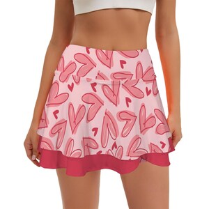 May include: A light pink athletic skirt featuring a repeating pattern of hand-drawn style hearts in darker pink and red outlines. The skirt has a high waistband and a solid dark pink ruffled hemline at the bottom, creating a layered appearance.