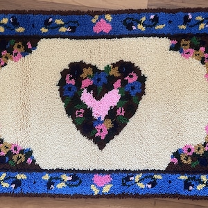 May include: A white rug with a brown and pink heart-shaped design in the center. The heart is surrounded by a floral pattern in shades of blue, pink, and green. The rug has a blue border with a brown and yellow floral pattern.