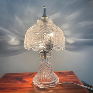 May include: A clear glass table lamp with a floral pattern on the shade and a diamond pattern on the base. The lamp has a silver-colored metal base and a gold-colored finial. The lamp is plugged into a wooden surface.