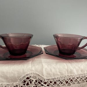 May include: Two matching vintage purple glass teacups and saucers. The cups have a triangular shape with a handle, and the saucers are hexagonal. The set is made of transparent glass, and the color is a deep, rich purple.
