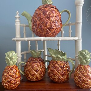 May include: A collection of ceramic pineapple-shaped tea and serving ware. The set includes a teapot, two lidded sugar bowls, a creamer, and a decorative pineapple. The pieces are brown with green accents.