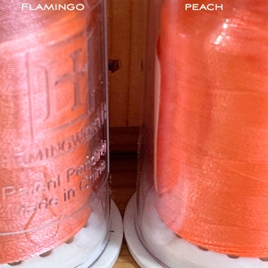 May include: Two spools of thread in clear plastic containers. The spool on the left is a light pink colour and labelled "Shy Flamingo". The spool on the right is a bright orange colour and labelled "Rebel Peach".