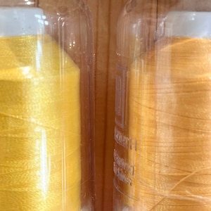 May include: Two spools of thread, one labelled "NEON YELLOW" and the other "MANGO". The thread on the left is a bright yellow, and the thread on the right is a more muted yellow.