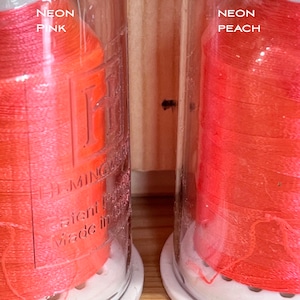 May include: Two spools of neon pink and neon peach embroidery floss in clear glass jars with white lids. The jars have the text "NEON PINK" and "NEON PEACH" printed on them. The spools are labelled "DMC" and "Made in Italy".