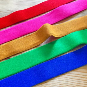 May include: Five different coloured elastic bands, red, pink, mustard yellow, green, and blue, are laid out on a wooden surface.