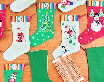 May include: A collection of colorful Christmas stockings with various festive designs, including Santa Claus, Christmas trees, and snowmen. The stockings are in shades of red, green, and blue, with numbers printed on the top. Wooden clothespins are also visible.
