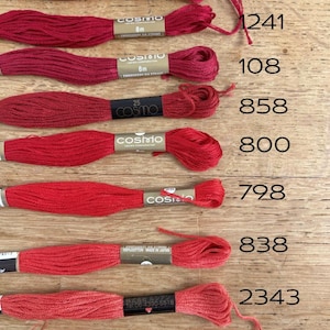May include: Nine spools of red embroidery floss with the brand name "Cosmo" printed on the labels. The spools are arranged vertically and labeled with numbers: 241A, 1241, 108, 858, 800, 798, 838, 2343, and 836.