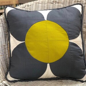 Puede incluir: Una almohada gris y blanca con un gran diseño de flor amarilla. La flor tiene un círculo en el centro y cuatro pétalos.
