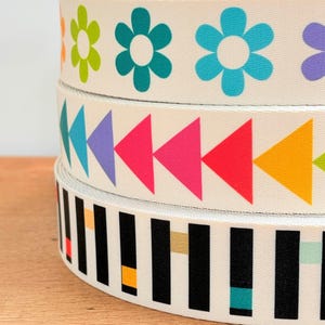 Peut inclure: Trois rouleaux de ruban en coton blanc avec différents motifs imprimés colorés. Le rouleau du haut présente un motif répétitif de fleurs vertes, turquoise et bleues. Le rouleau du milieu présente un motif répétitif de triangles violets, roses, rouges et jaunes. Le rouleau du bas présente un motif répétitif de rayures noires et blanches avec des accents de jaune, orange et turquoise.