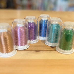 May include: Six spools of metallic thread in various colours, including gold, pink, purple, blue, teal, and green, are displayed in clear glass jars with white lids. The spools are arranged in a row on a wooden surface.