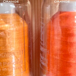 May include: Two spools of thread in clear glass containers. The spool on the left is labelled "TROPICAL ORANGE" and the spool on the right is labelled "NEON ORANGE".