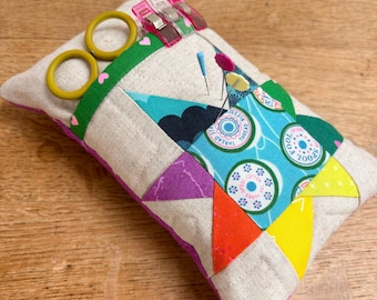 May include: A rectangular fabric pincushion featuring a patchwork design with beige, green, blue, orange, and yellow sections. It includes three pink sewing clips, a pair of small yellow scissors, and several sewing pins inserted into the top.