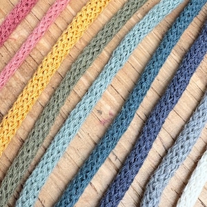 Può includere: Primo piano di diversi fili di corda intrecciata in vari colori, tra cui rosa, giallo, verde, blu e bianco. I cordoncini sono disposti diagonalmente su una superficie di legno marrone chiaro, mostrando la loro consistenza e le variazioni di colore.