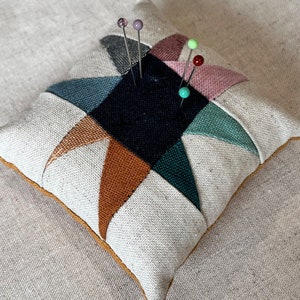 Può includere: Un cuscino per spilli in tessuto bianco con un design a stella geometrico in tonalità di marrone, verde e rosa. Il cuscino per spilli ha un bordo dorato ed è pieno di spilli con teste di vetro colorate.