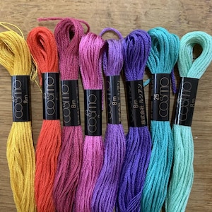 May include: A collection of eight skeins of Cosmo embroidery floss in a variety of colors, including yellow, orange, pink, purple, teal, and green. Each skein is labeled with the Cosmo brand name and 8m length.