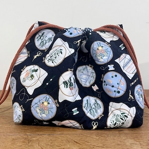 以下が含まれることがあります： 刺繍枠に花やサボテンのデザインが描かれた、キルティングされたデザインの黒い巾着袋。袋には茶色の紐が付いています。