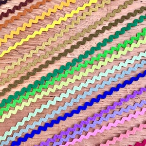 May include: A collection of 20 different coloured zig zag trim, each trim is a different colour, ranging from dark brown to red. The trim is arranged in a diagonal pattern on a wooden surface.