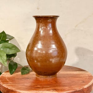May include: A brown ceramic vase with a rounded body and a narrow neck. The vase has a glossy finish and sits on a wooden, round pedestal. The pedestal is approximately 12 inches in diameter and has three small legs.