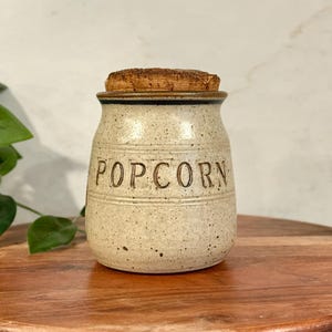 May include: A speckled ceramic popcorn jar with a cork lid. The jar has the word "POPCORN" embossed on the front. The jar is sitting on a wooden surface.