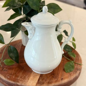 May include: A white ceramic coffee pot with a lid and handle. The pot is on a wooden serving board, with green plants in the background. The pot has a decorative spout and handle.