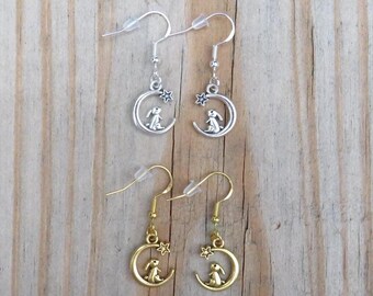 May include: A pair of silver and gold earrings featuring a crescent moon with a small star and a rabbit sitting on the moon.