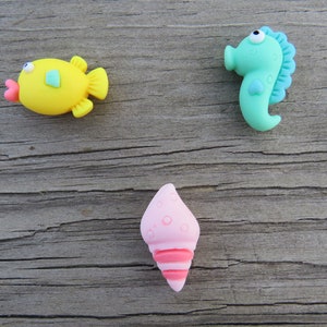 May include: Three colorful sea-themed embellishments: a yellow fish with a pink mouth, a turquoise seahorse, and a pink seashell with red stripes. These decorative items are perfect for craft projects.