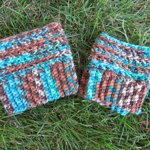 May include: A pair of hand-knitted wrist warmers in shades of brown, teal, and white. The textured knitwear features a ribbed cuff and a patterned design, perfect for adding warmth and style. The wrist warmers are displayed on a bed of green grass.