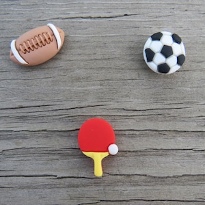 May include: Three miniature sports-themed figurines: a brown and white football, a black and white soccer ball, and a red and yellow ping pong paddle with a white ball.