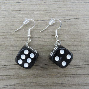 May include: A pair of black dice earrings with white dots. The earrings have silver-colored hooks and clear plastic ear protectors. The dice are square-shaped and have a glossy finish. The earrings are displayed on a wooden surface.