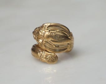 May include: A gold-colored ring with an ornate, swirling design. The ring appears to be made from a spoon, with the spoon's handle wrapping around the finger. The ring is set against a white background.