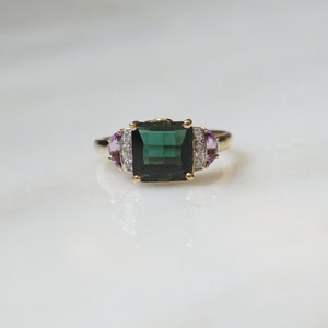Pu&ograve; includere: Un anello d'oro con una grande pietra preziosa verde smeraldo circondata da pietre preziose pi&ugrave; piccole, chiare e viola.