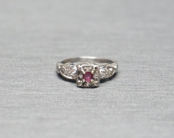 May include: A silver ring with a square, red gemstone at the center. The ring has ornate detailing on the band and around the gemstone. The ring is set against a gray background.