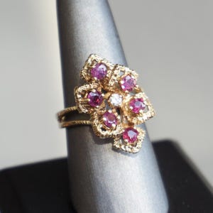 May include: A gold-toned ring featuring a floral design with multiple round, deep red gemstones and a single, smaller white gemstone at the center. The ring is displayed on a gray ring holder.