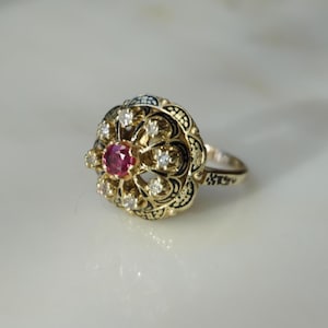 May include: A gold-toned ring with a central red gemstone, surrounded by smaller clear stones. The ring has an ornate, floral-inspired design with black detailing. The ring is set against a white background.