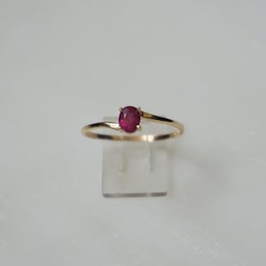 May include: A gold-toned ring featuring a vibrant red oval gemstone. The band gracefully curves around the finger, with the gemstone set in a delicate, minimalist design. The ring is displayed on a clear stand against a white background.