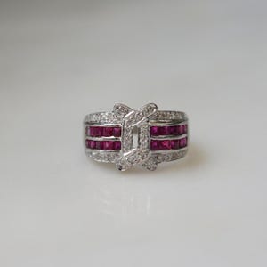 May include: A silver-toned ring featuring two horizontal rows of square-cut deep red rubies set in a channel. The center of the band is accented by a vertical, overlapping crossover design encrusted with small, clear, round-cut white stones.